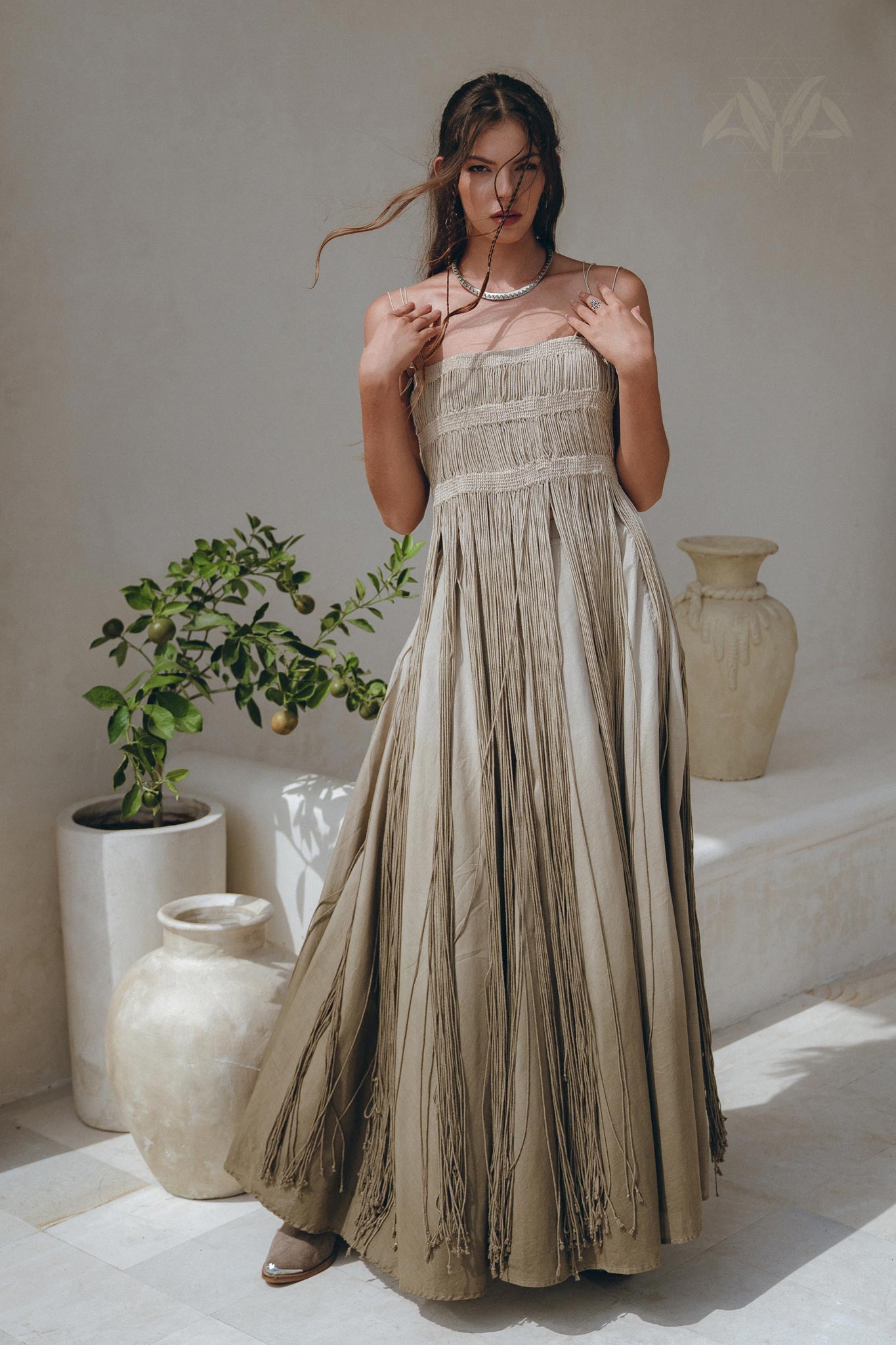 A woman in the Sage Green Tribal Goddess Slip Over by AYA Sacred Wear—an ethnic handwoven macrame layer-up sheer net dress—stands indoors by large ceramic vases and a green plant, her hair loose, one hand touching her necklace and dress.