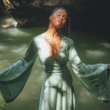 A model poses gracefully in a serene setting, wearing a sky blue goddess dress from Aya Sacred Wear. The dress features flowing sleeves and a deep V-neckline, with soft light filtering through the trees, creating an ethereal atmosphere.