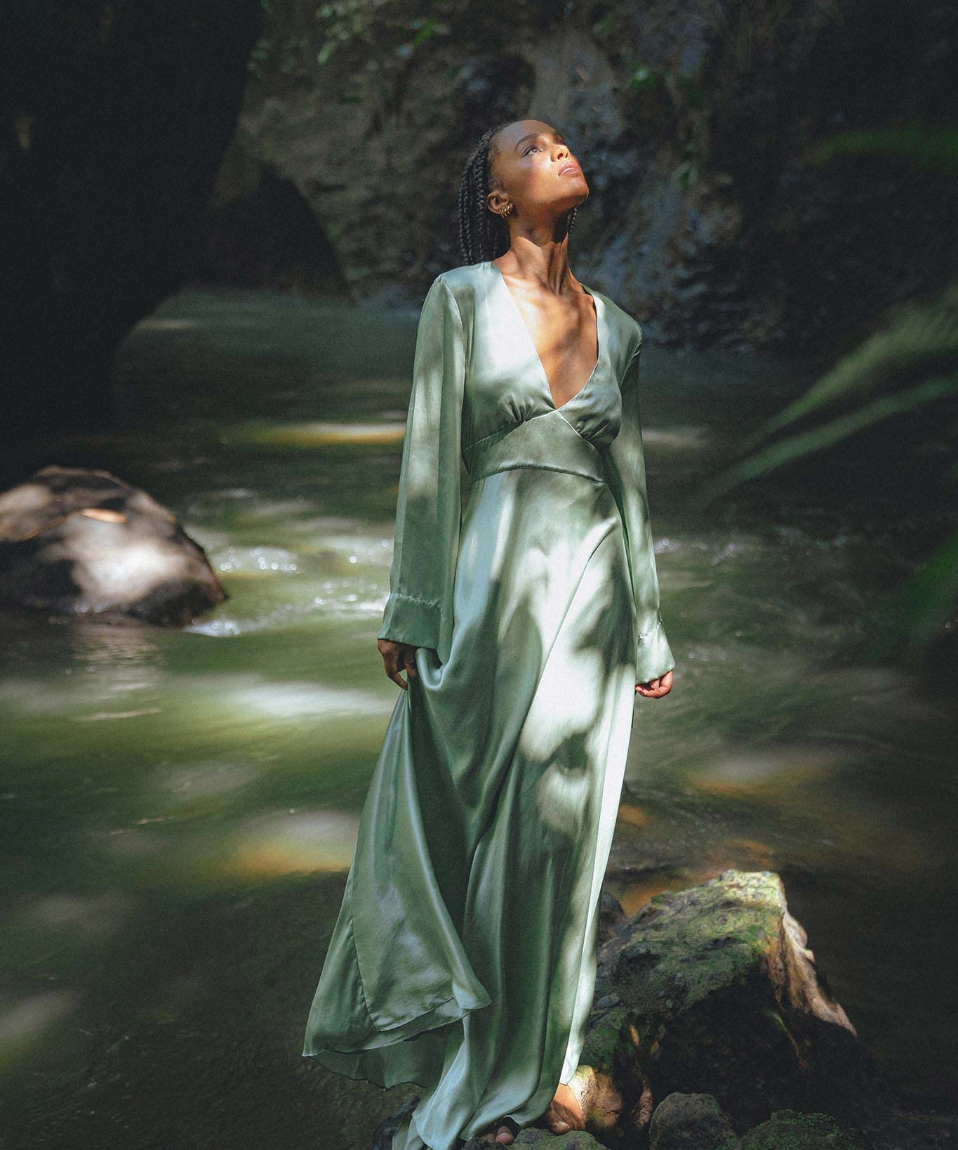 A woman elegantly poses in a sky blue goddess dress, designed by Aya Sacred Wear, perfect for a bridesmaid. The dress features flowing fabric with delicate draping that cascades gracefully. The backdrop shows a picturesque outdoor setting with greenery and soft lighting, enhancing the ethereal quality of the gown.