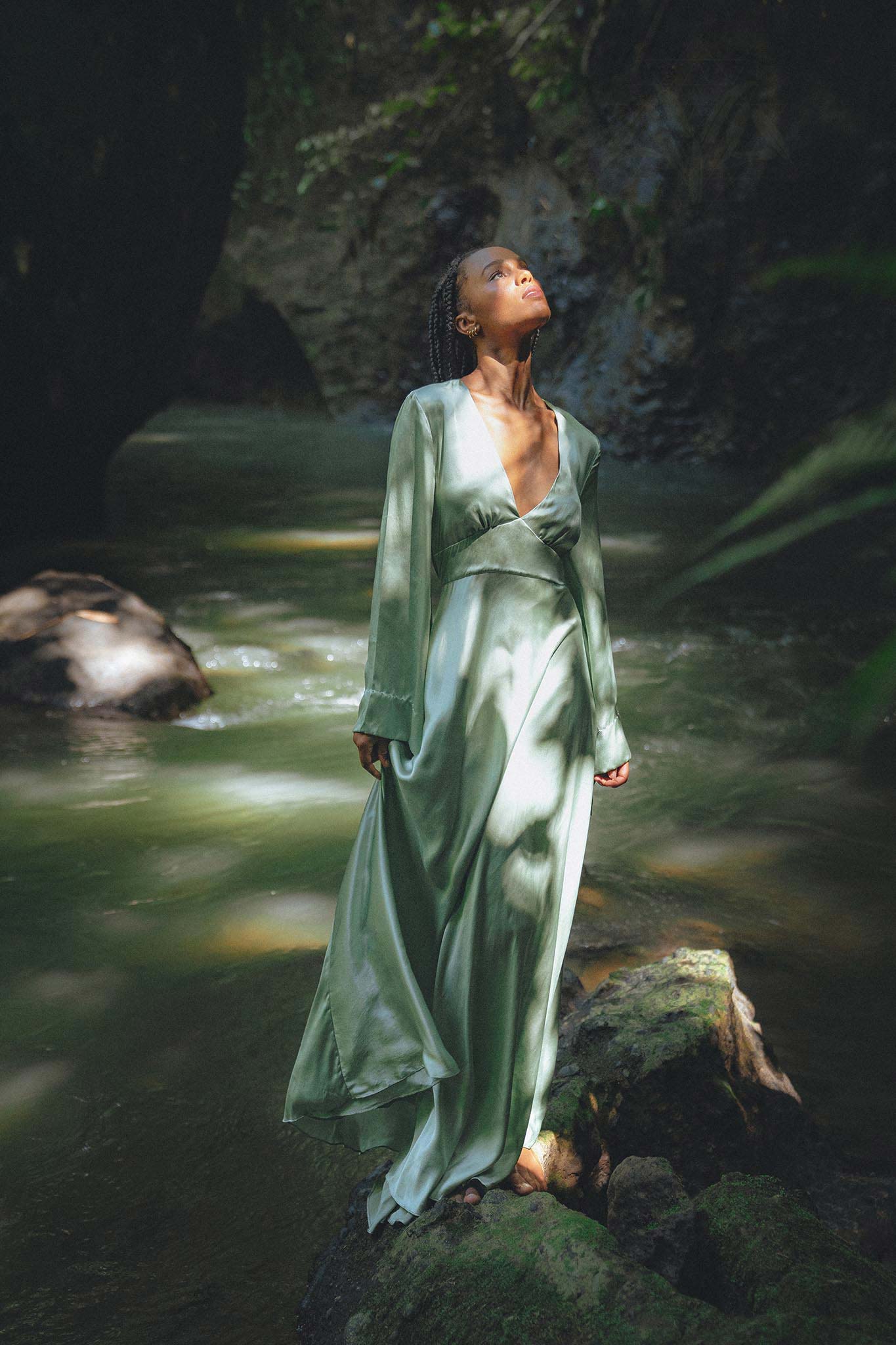 A woman elegantly poses in a sky blue goddess dress, designed by Aya Sacred Wear, perfect for a bridesmaid. The dress features flowing fabric with delicate draping that cascades gracefully. The backdrop shows a picturesque outdoor setting with greenery and soft lighting, enhancing the ethereal quality of the gown.
