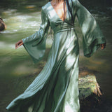 A woman with long braids is gracefully standing in a flowing dress with bell sleeves, surrounded by a serene natural setting. The soft fabric of the dress catches the sunlight, creating a magical effect. She is positioned on rocks near a gentle stream, embodying an ethereal vibe. This is the sky blue goddess dress, designed as a bridesmaid dress by Aya Sacred Wear.