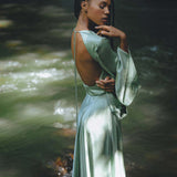 A woman poses gracefully in a backless, sky blue goddess dress by Aya Sacred Wear, standing near a serene water flow. The dress features long, flowing sleeves and soft drapes that enhance her elegance. Sunlight filters through the trees, creating a beautiful interplay of light and shadow on her skin and the dress.