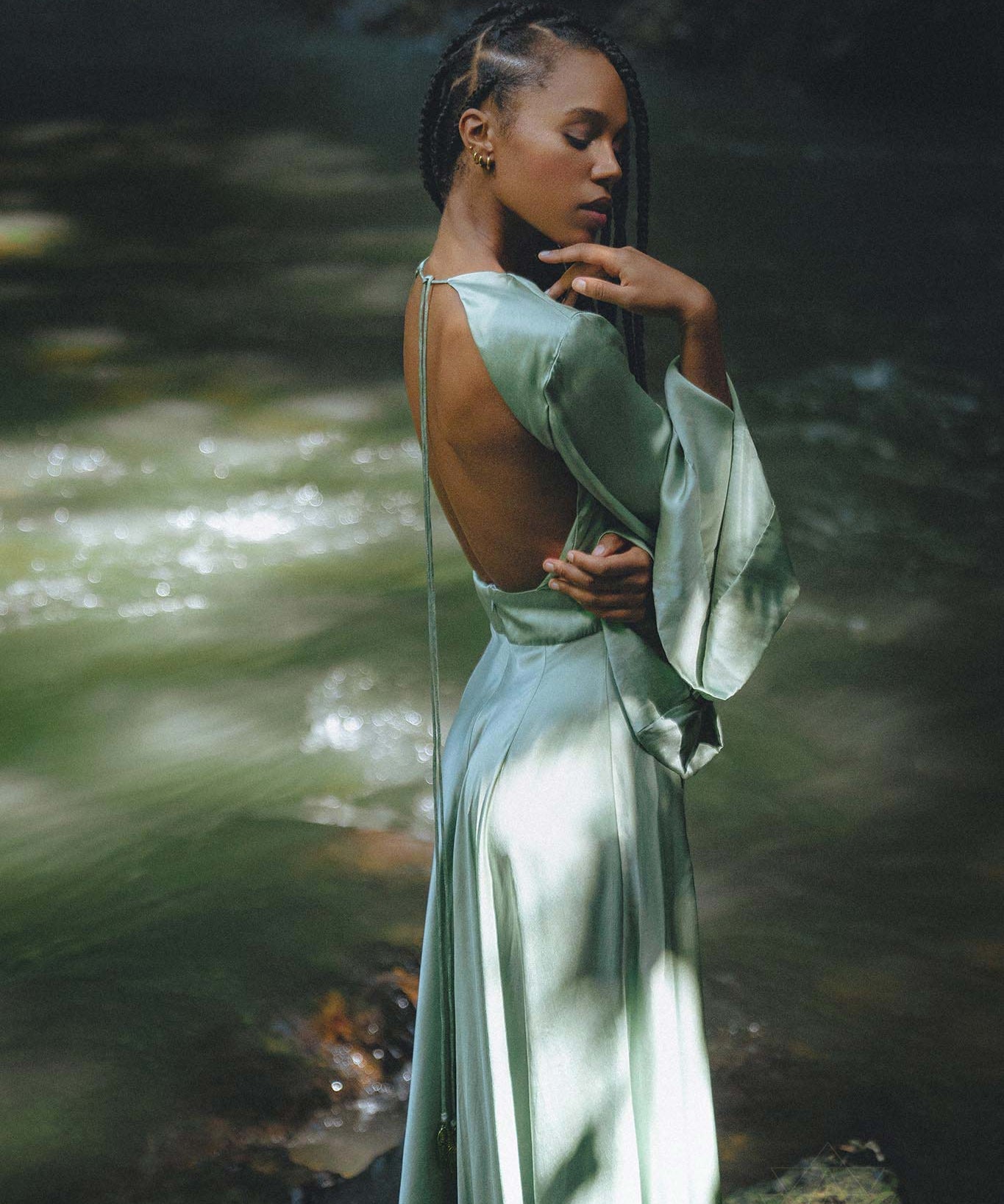 A woman poses gracefully in a backless, sky blue goddess dress by Aya Sacred Wear, standing near a serene water flow. The dress features long, flowing sleeves and soft drapes that enhance her elegance. Sunlight filters through the trees, creating a beautiful interplay of light and shadow on her skin and the dress.