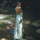 A woman stands on a mossy rock by a serene river, wearing a beautiful sky blue goddess dress. The dress features an elegant back design and flowing fabric that catches the soft sunlight filtering through the trees. This enchanting look embodies the aesthetic of a bridesmaid dress by Aya Sacred Wear.