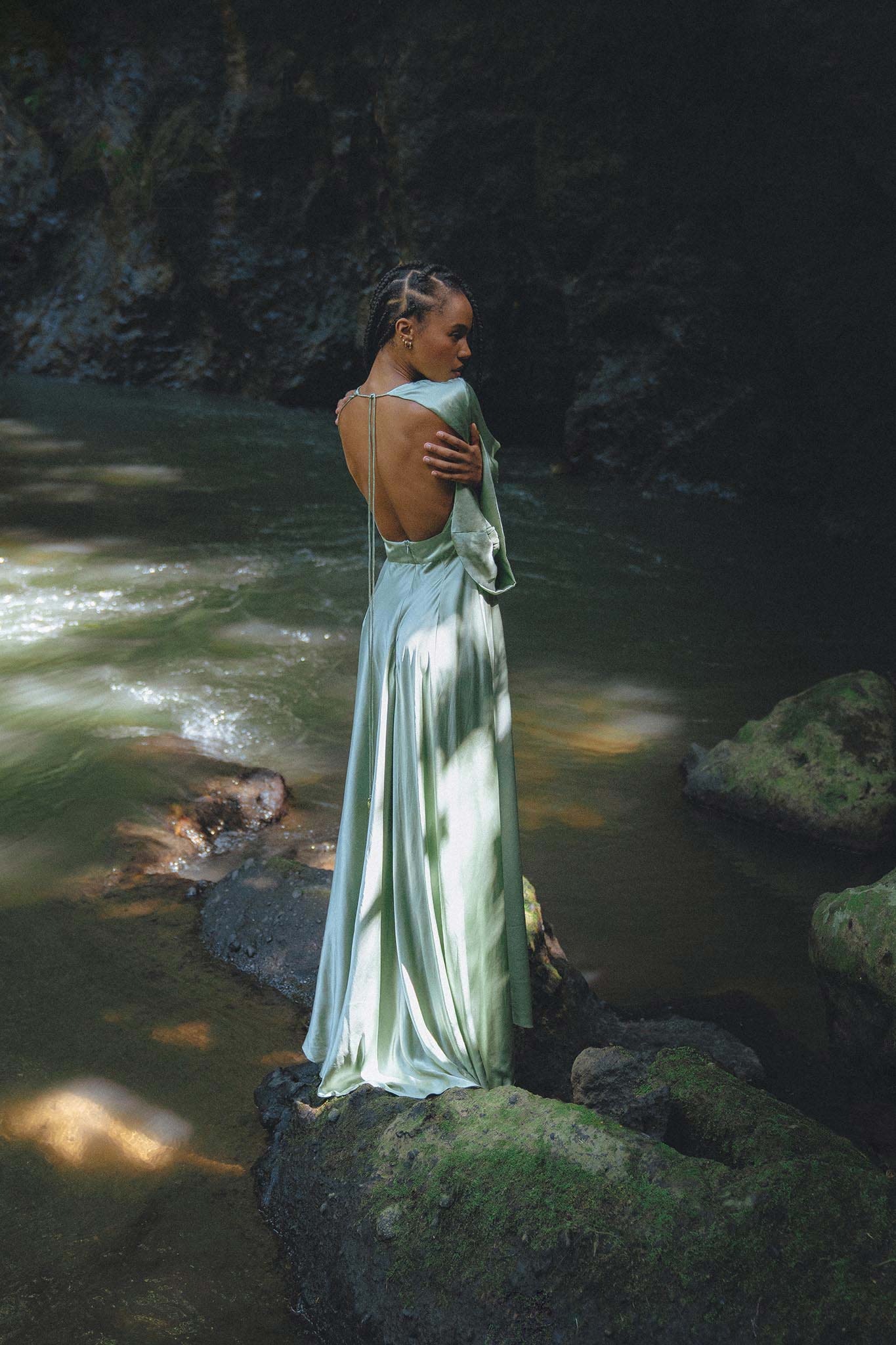 A woman stands on a mossy rock by a serene river, wearing a beautiful sky blue goddess dress. The dress features an elegant back design and flowing fabric that catches the soft sunlight filtering through the trees. This enchanting look embodies the aesthetic of a bridesmaid dress by Aya Sacred Wear.