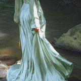 A model wearing a flowing, sky blue goddess dress from Aya Sacred Wear stands by a serene water setting, showcasing the graceful back and long sleeves of the garment as the fabric drapes elegantly around her. The dress catches the light, highlighting its silky texture and movement.