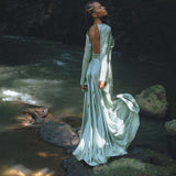 A woman stands gracefully on a rock by the water, wearing a stunning sky blue goddess dress from Aya Sacred Wear. The dress flows elegantly, showcasing a backless design and long sleeves. The lush background features a dark, rocky terrain, while the sunlight casts a gentle glow on the fabric and surrounding nature, creating a serene and ethereal atmosphere.