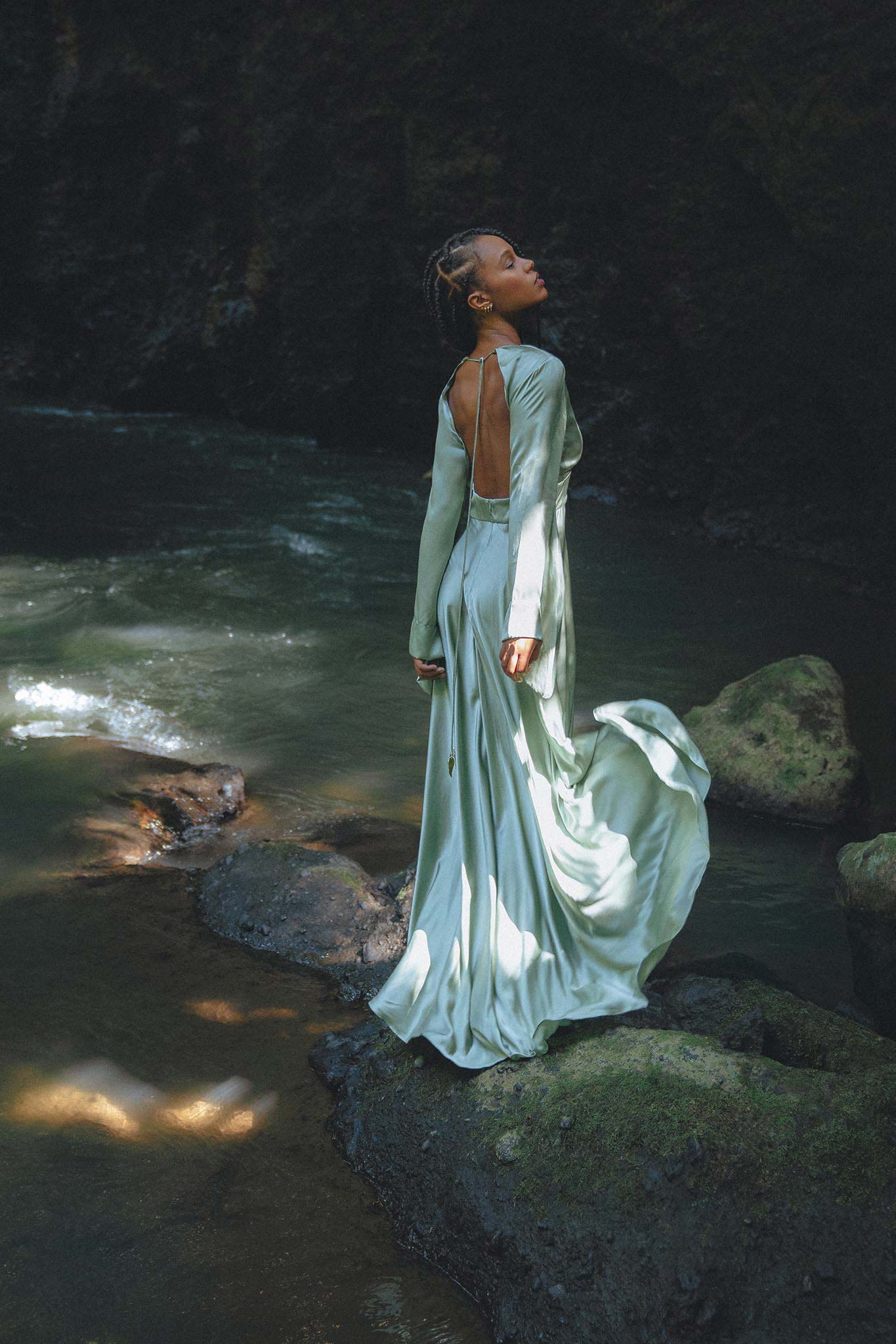 A woman stands gracefully on a rock by the water, wearing a stunning sky blue goddess dress from Aya Sacred Wear. The dress flows elegantly, showcasing a backless design and long sleeves. The lush background features a dark, rocky terrain, while the sunlight casts a gentle glow on the fabric and surrounding nature, creating a serene and ethereal atmosphere.