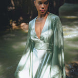A person stands gracefully near a serene body of water, wearing a flowing dress with long sleeves and a deep V-neckline. The dress, in a soft mint green shade, complements the natural surroundings. Sunlight filters through the trees, casting gentle shadows on the dress, enhancing its elegant silhouette. This outfit is part of the "Sky Blue Goddess Dress" collection, designed by Aya Sacred Wear.