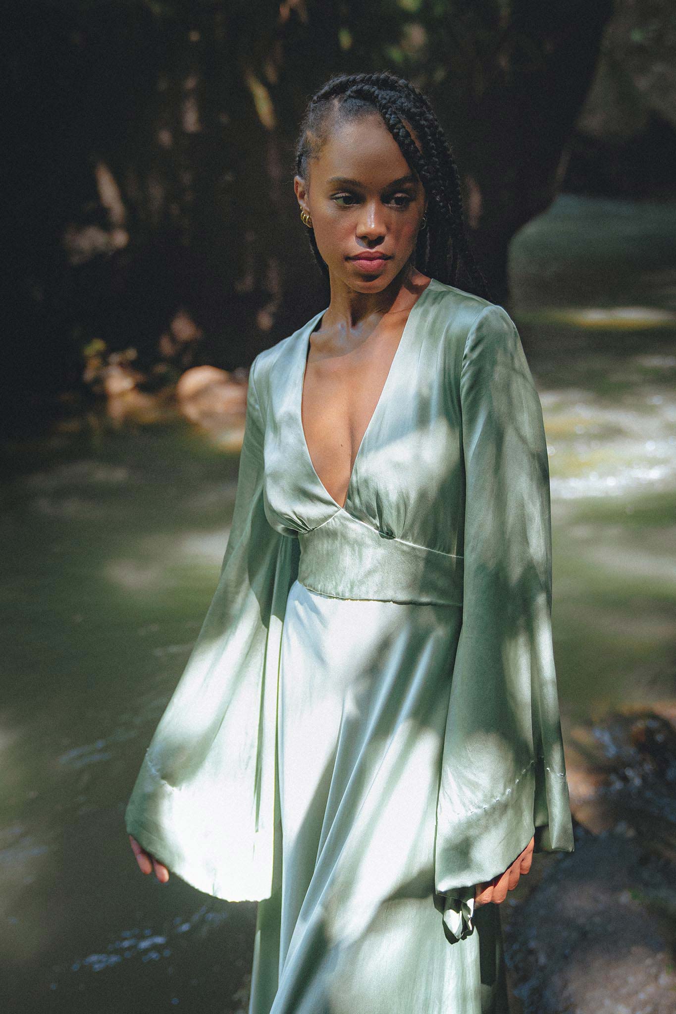A person stands gracefully near a serene body of water, wearing a flowing dress with long sleeves and a deep V-neckline. The dress, in a soft mint green shade, complements the natural surroundings. Sunlight filters through the trees, casting gentle shadows on the dress, enhancing its elegant silhouette. This outfit is part of the "Sky Blue Goddess Dress" collection, designed by Aya Sacred Wear.