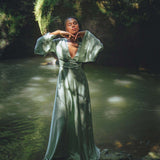 A model poses gracefully in a serene natural setting, wearing a sky blue goddess dress designed by Aya Sacred Wear. The dress features flowing sleeves and a deep V neckline, complementing the tranquil atmosphere of the water and lush greenery surrounding her.