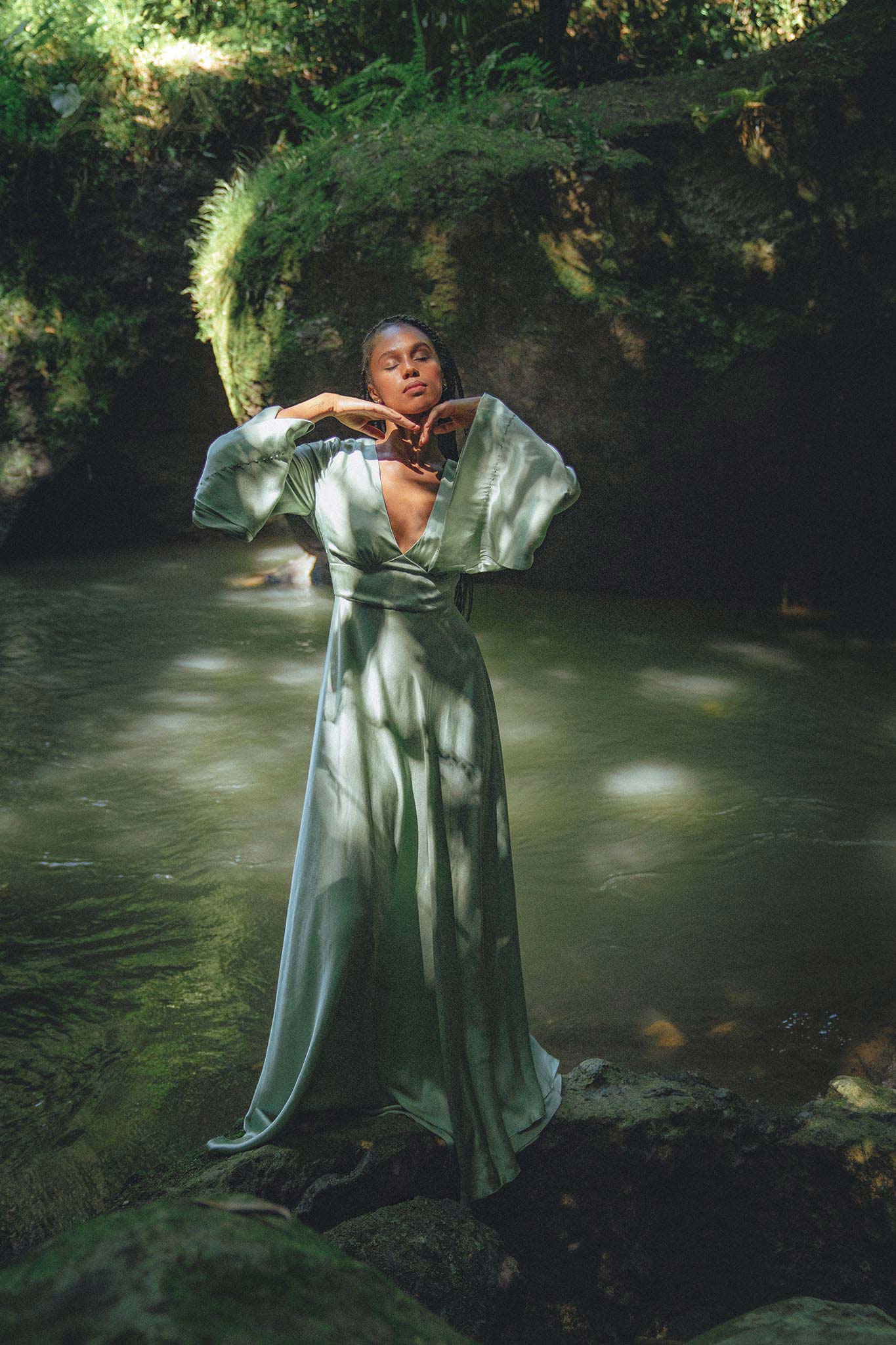 A model poses gracefully in a serene natural setting, wearing a sky blue goddess dress designed by Aya Sacred Wear. The dress features flowing sleeves and a deep V neckline, complementing the tranquil atmosphere of the water and lush greenery surrounding her.