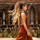 A woman with long hair in a ponytail, wearing an AYA Sacred Wear Red Boho Bridesmaid Dress, walks past brick arches and statues. Tall grass sways gently in the foreground, adding movement and tranquility to the scene.