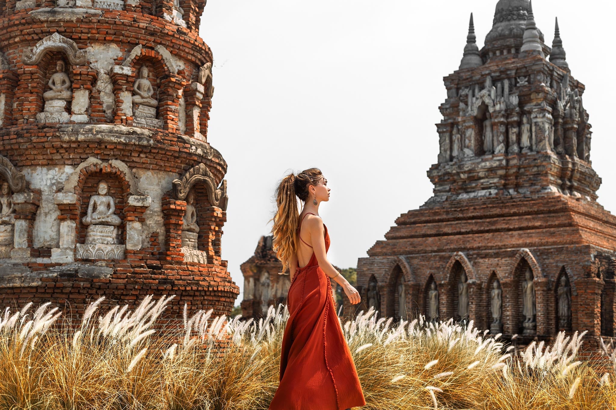 A woman wearing an AYA Sacred Wear Red Boho Bridesmaid Dress strolls through tall grass before ancient brick temple ruins with intricate carvings and statues under a clear sky.