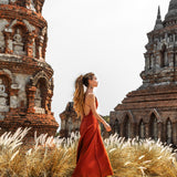 A woman wearing an AYA Sacred Wear Red Boho Bridesmaid Dress strolls through tall grass before ancient brick temple ruins with intricate carvings and statues under a clear sky.