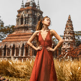 Wearing the handmade Goddess V Neck Dress by AYA Sacred Wear, a woman confidently stands with hands on hips in front of an ancient brick temple with ornate spires. Tall grass surrounds the area beneath a clear sky.