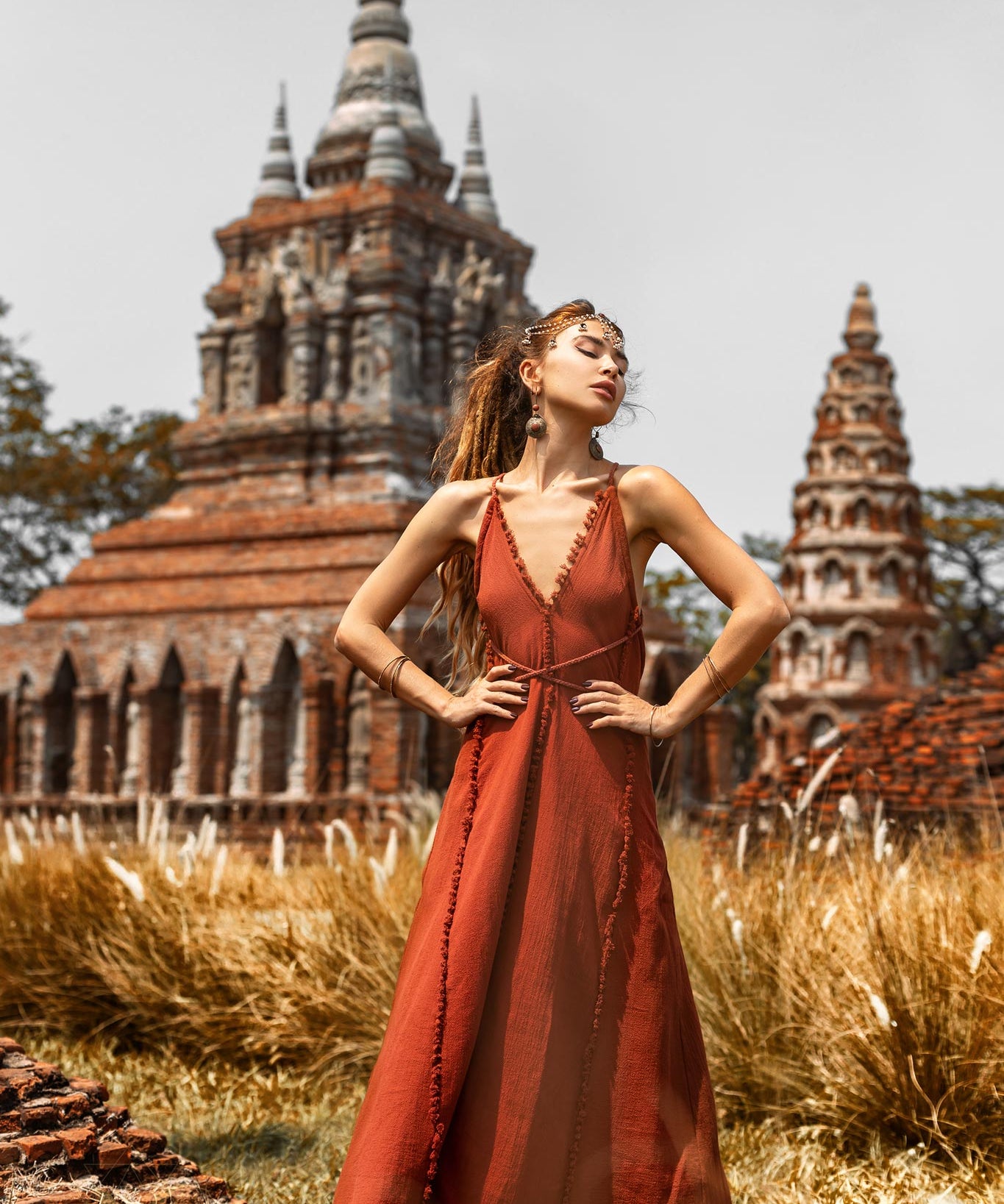 Wearing the handmade Goddess V Neck Dress by AYA Sacred Wear, a woman confidently stands with hands on hips in front of an ancient brick temple with ornate spires. Tall grass surrounds the area beneath a clear sky.