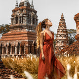 A woman in an AYA Sacred Wear Red Boho Bridesmaid Dress poses gracefully in a grassy area, with ancient brick pagodas behind her, exuding historical exploration and tranquility.