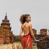 Dressed in AYA Sacred Wear's Red Boho Bridesmaid Dress, a woman stands in a field of tall grass, gazing at an ancient brick structure with a pagoda-like tower. The overcast sky enhances the mysterious aura of this historical site, evoking another era.