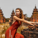 A woman in an AYA Sacred Wear Red Boho Bridesmaid Dress leans on a brick wall in a field, framed by ancient temples. The scene is serene, with tall grass and an overcast sky. Her long hair flows freely, adorned with jewelry that complements the handmade fabric of her attire.
