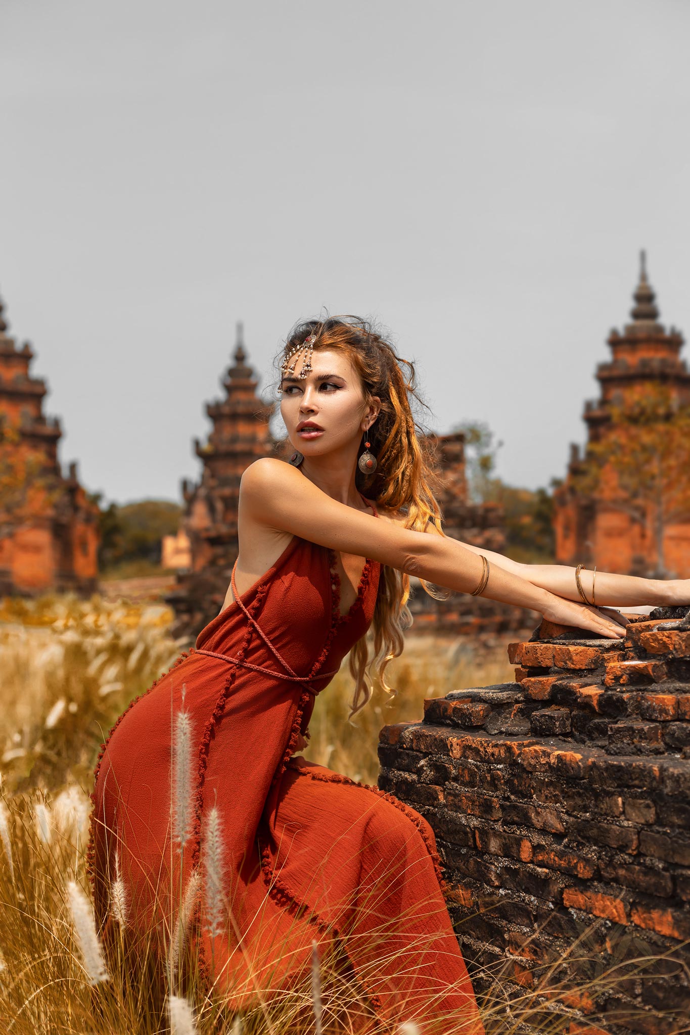 A woman in an AYA Sacred Wear Red Boho Bridesmaid Dress leans on a brick wall in a field, framed by ancient temples. The scene is serene, with tall grass and an overcast sky. Her long hair flows freely, adorned with jewelry that complements the handmade fabric of her attire.