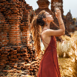 Wearing the Red Boho Bridesmaid Dress by AYA Sacred Wear, a woman stands among ancient brick ruins. The backless, flowing design complements her bohemian braids as she gazes upward with hands raised. The sun casts a warm glow, highlighting tall grasses in the background.