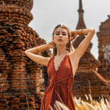 A woman poses against ancient brick structures in a Red Boho Bridesmaid Dress by AYA Sacred Wear, her hands behind her head, wearing a headpiece and bracelets. Standing amidst tall grass under a cloudy sky, she exudes an effortlessly chic vibe.