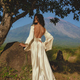 A woman in a Vanilla Cream Sundari Silk Gown by AYA Sacred Wear stands under a tree, facing away. She overlooks a green field with a distant mountain under the clear blue sky, embodying elegance and serenity.
