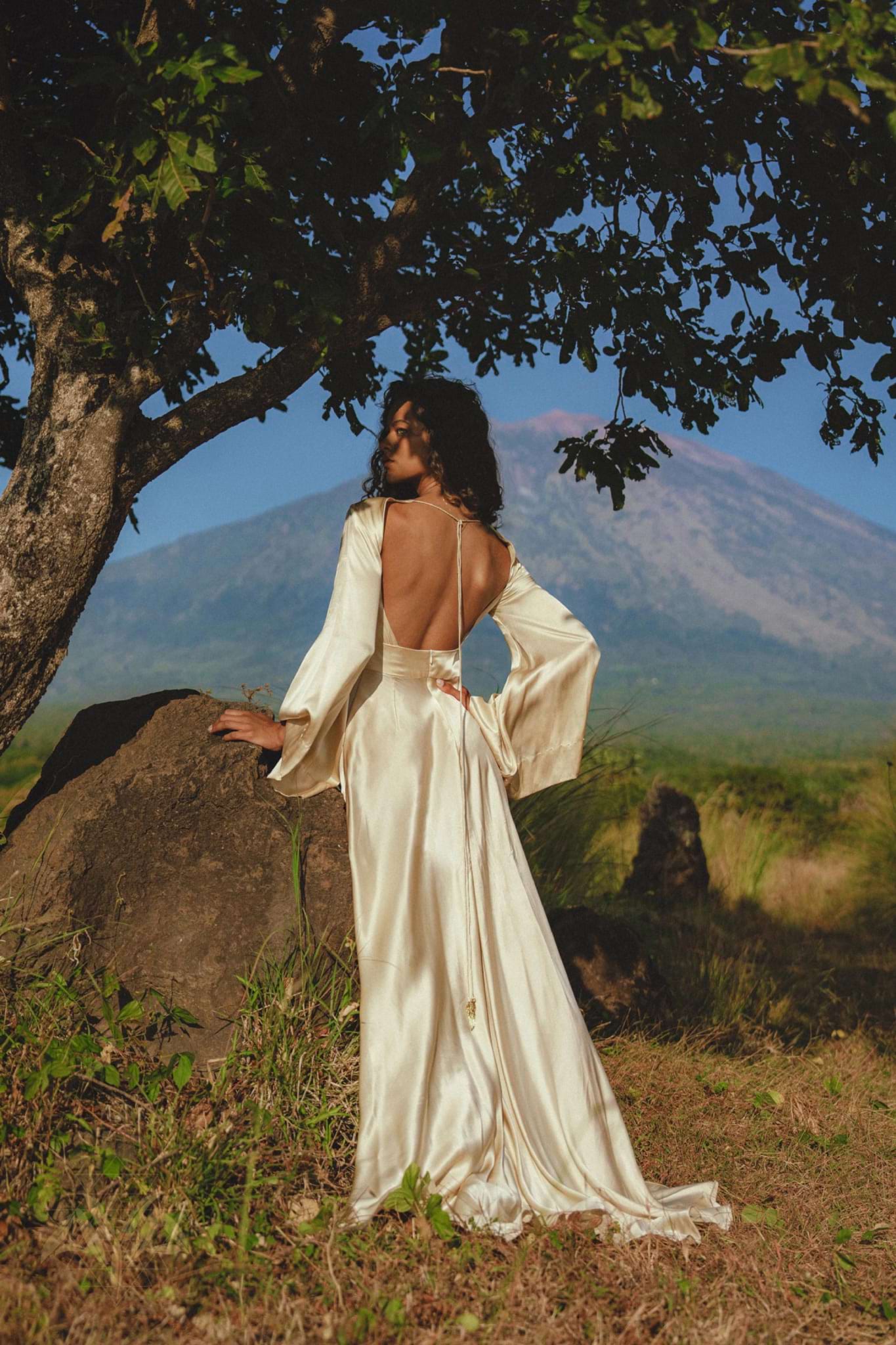 A woman in a Vanilla Cream Sundari Silk Gown by AYA Sacred Wear stands under a tree, facing away. She overlooks a green field with a distant mountain under the clear blue sky, embodying elegance and serenity.