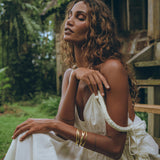 A woman with curly hair sits outside near a rustic wooden structure, wearing the Boho Beach Macrame Wedding Dress by AYA Sacred Wear. This off-white, multiway bohemian dress complements her relaxed demeanor as she closes her eyes. Gold bracelets adorn her wrists amid the lush green foliage that surrounds the area.
