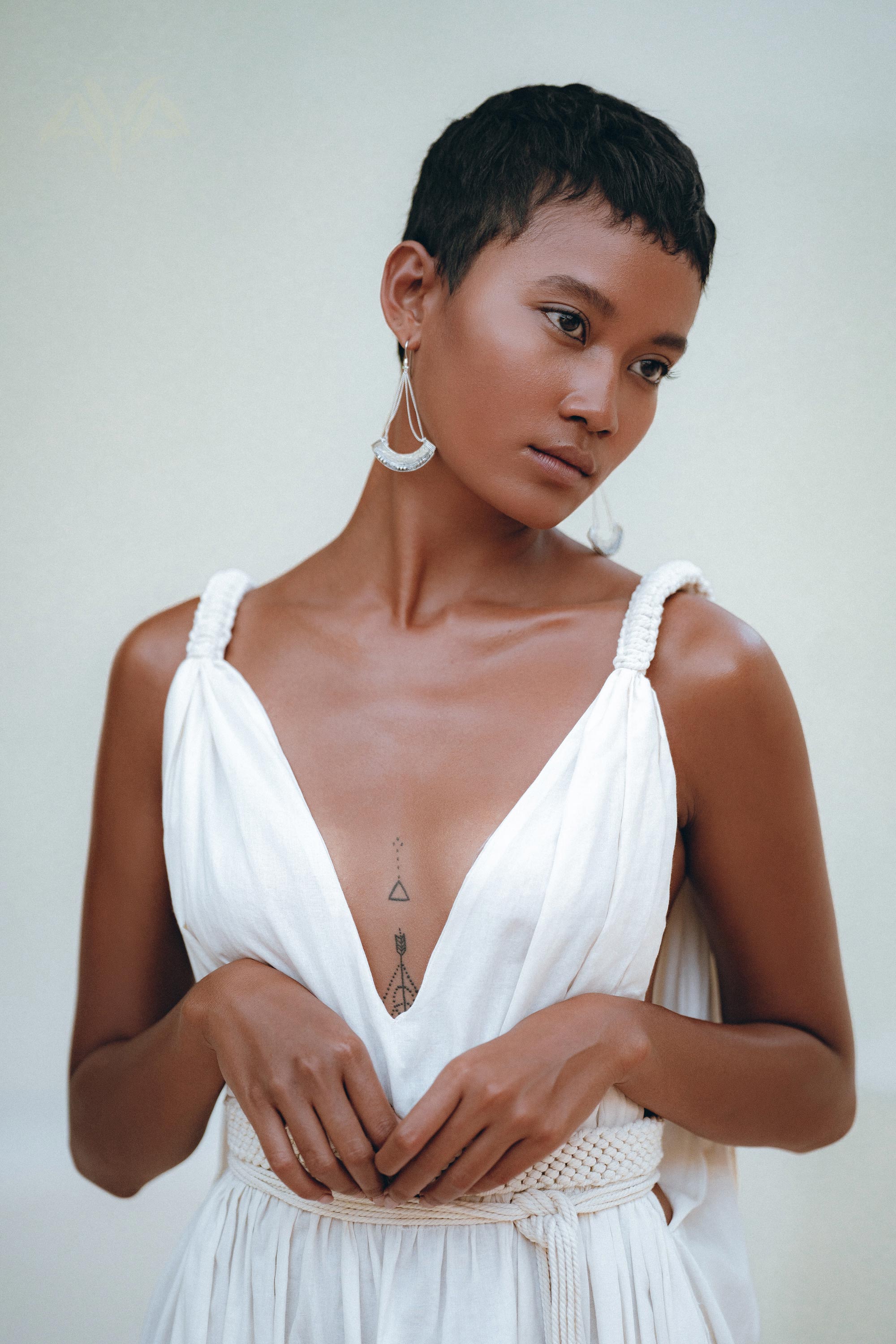 A person with short hair is seen standing against a neutral background, wearing the Boho Beach Macrame Wedding Dress by AYA Sacred Wear. The off-white multiway bohemian dress showcases their geometric chest tattoos as they gaze to the side, with one hand clasping the other. An artisanal macrame belted waist completes this unique look.
