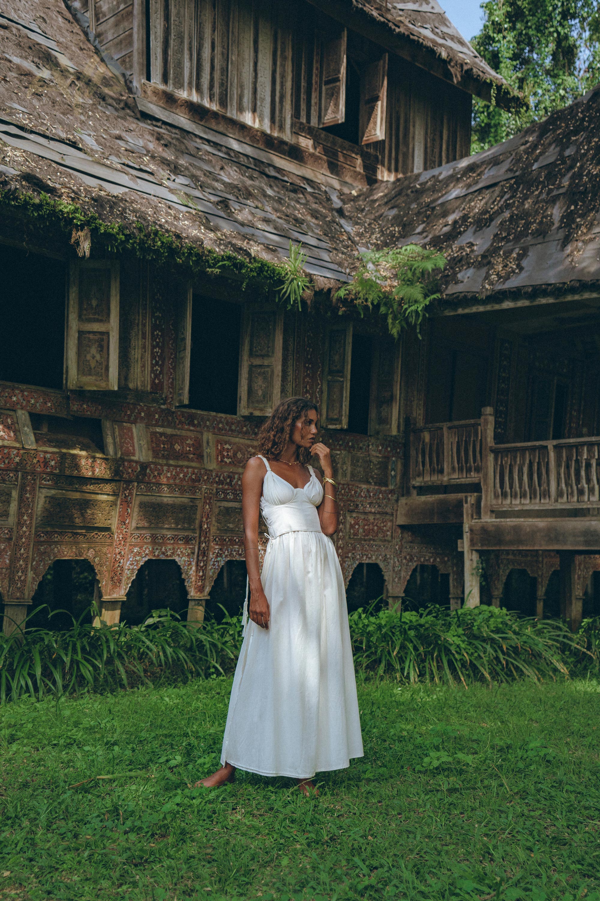 Dressed in an AYA Sacred Wear Boho Beach Macrame Wedding Dress, an Off-White Multiway Bohemian piece, a woman gazes thoughtfully in front of a traditional wooden house adorned with intricate carvings and lush greenery. This aged structure, resembling a unique piece of art, features a weathered roof and decorative patterns on its walls.