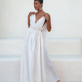 Against a minimalist backdrop, a person poses gracefully in the Boho Beach Macrame Wedding Dress by AYA Sacred Wear. This off-white, multiway bohemian dress features a flowing, sleeveless design crafted from light homespun cotton fabric with a deep V-neckline and cinched waist. Paired with white sandals and large hoop earrings, they exude the enchanting charm of this boho-inspired attire.