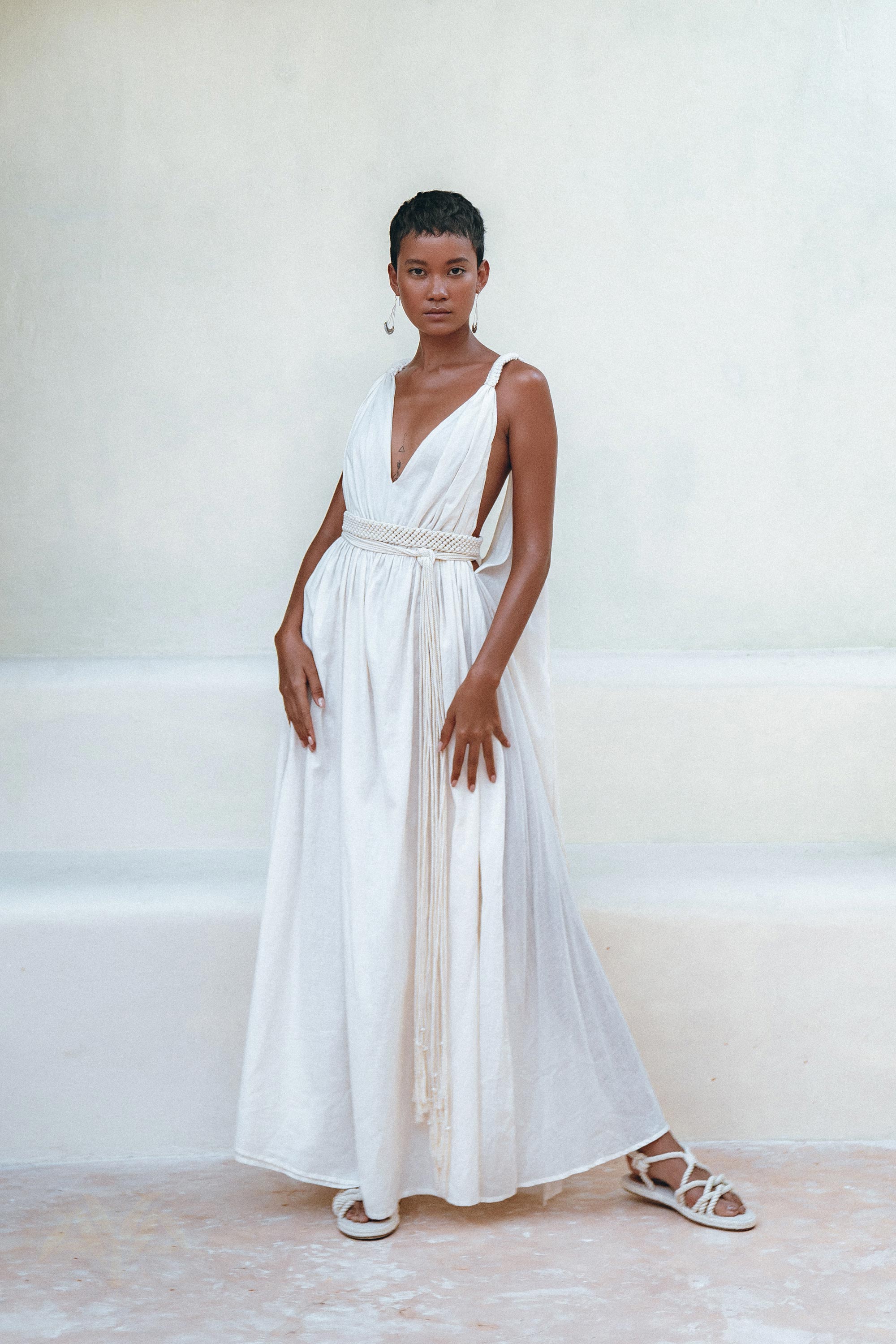 Against a light background, a confident individual with short hair showcases the elegance of the AYA Sacred Wear's Boho Beach Macrame Wedding Dress. Featuring a deep V-neck and tied waist, this off-white multiway bohemian dress is crafted with homespun cotton, perfectly accessorized with hoop earrings, macrame belted details, and white sandals.