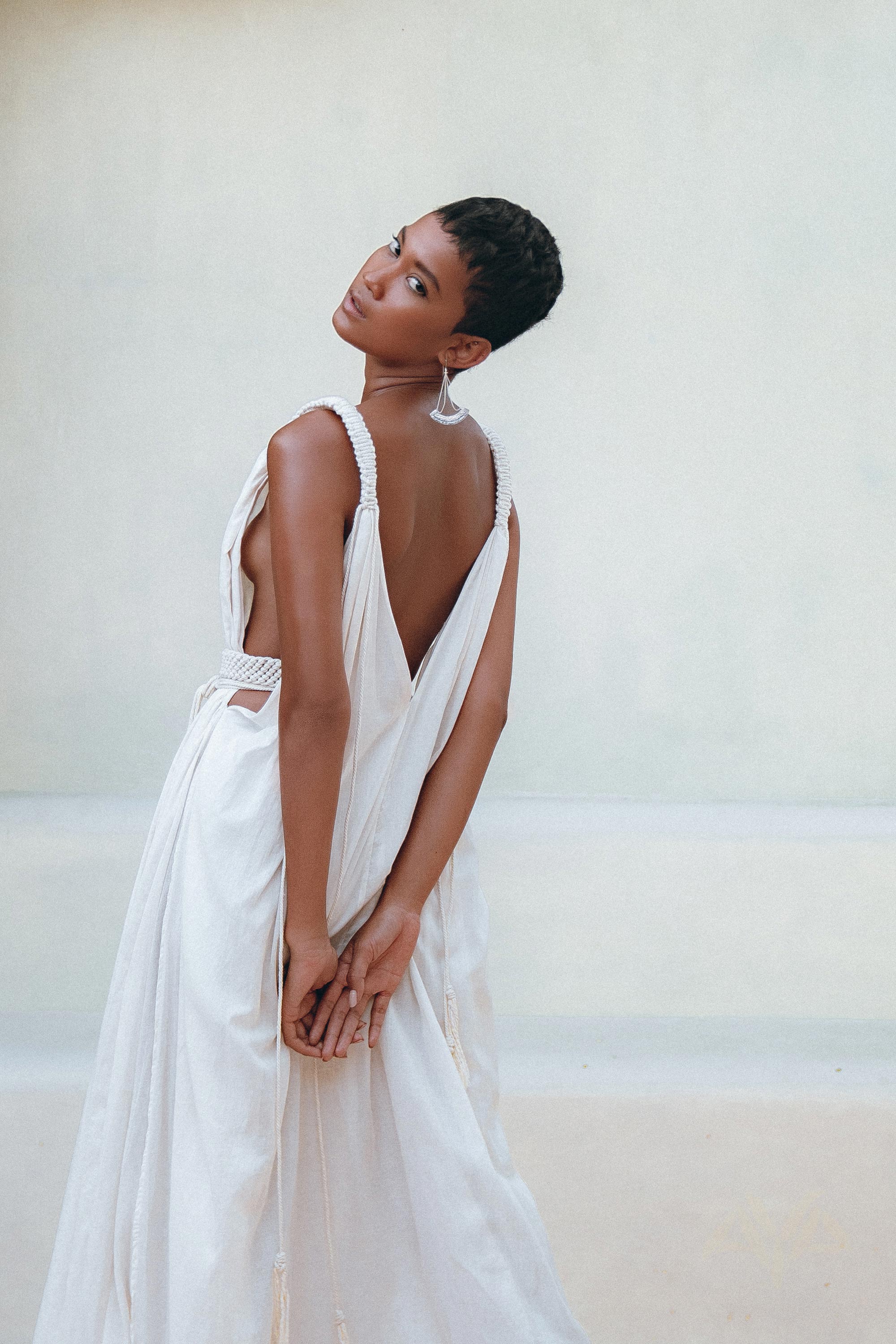 Wearing the Boho Beach Macrame Wedding Dress by AYA Sacred Wear, featuring an off-white multiway design, a person stands gracefully with their head tilted and hands clasped behind. The elegant bohemian dress beautifully complements the neutral outdoor backdrop, creating a scene reminiscent of a living piece of art.