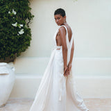 Wearing the Boho Beach Macrame Wedding Dress by AYA Sacred Wear, an individual stands gracefully before a cream-colored wall adorned with vibrant green foliage. With a calm expression, they glance over their shoulder. Nearby, a large white ceramic pot adds an artistic touch to the scene.